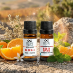 Fotografía de alta gama de dos aceites esenciales de naranja y menta de Cuatro Cuatro sobre piedra rústica, rodeados de rodajas de naranja, hojas de menta frescas y destellos dorados en el paisaje natural de Ensenada.